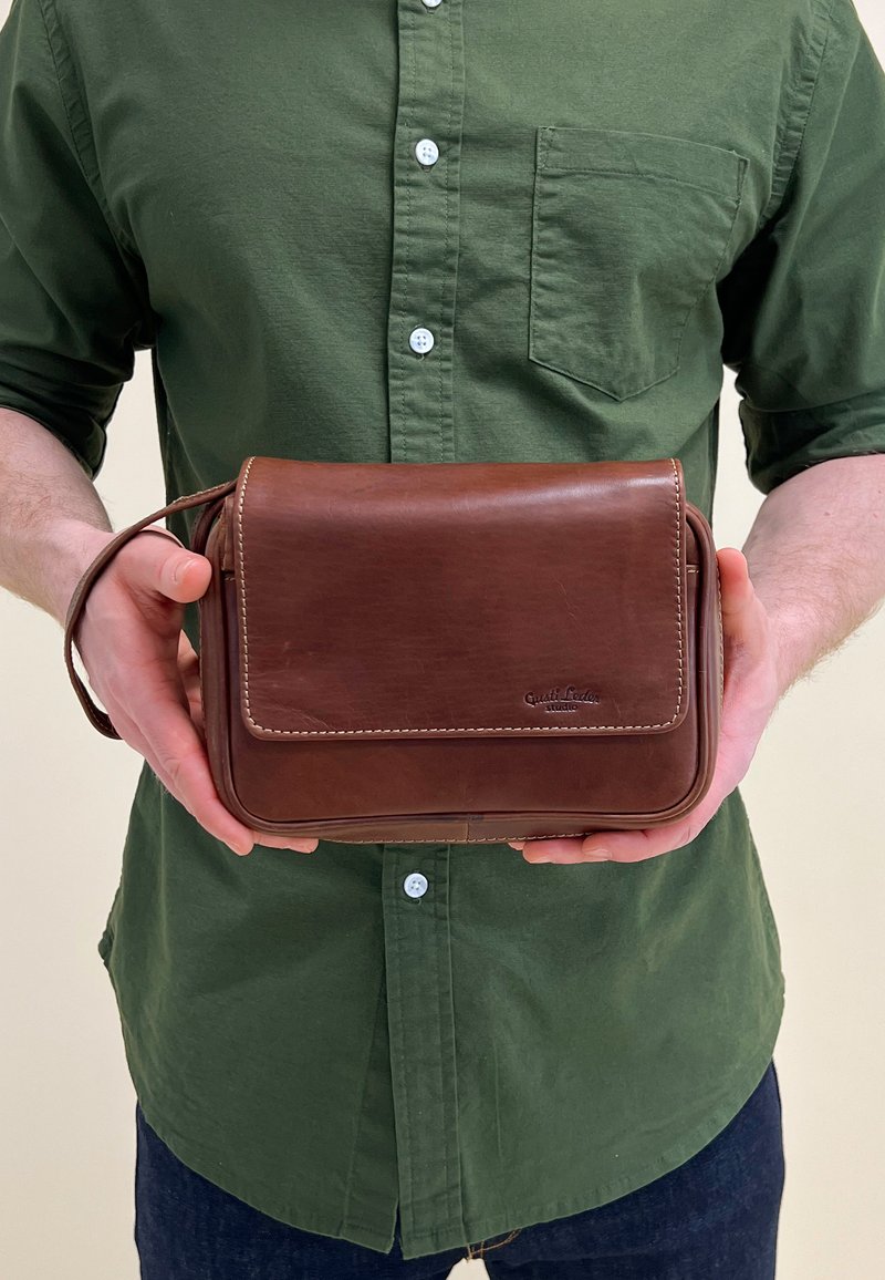 Brun läder crossbody-väska med klaff, vit sömnad och sidohandtag. Rektangulär form, slät textur, visar logotypdetalj på framsidan.