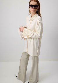 Chemise en soie crème avec un col, des manches évasées et des poignets texturés ; associée à un pantalon large gris clair et des chaussures noires à talons pointus.