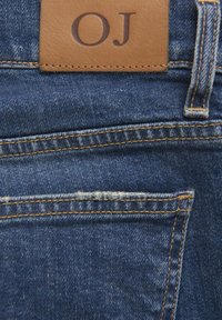 Denim w ciemnoniebieskim odcieniu, z brązową skórzaną łatą z napisem "OJ", z widocznym przeszyciem i małym detalem tylnej kieszeni.