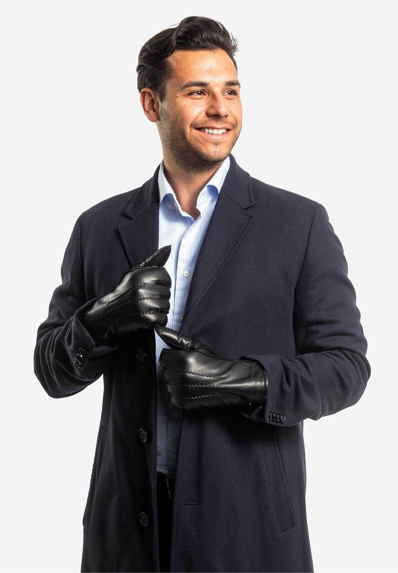 Schwarze Lederhandschuhe mit strukturierten Nähten, getragen von einem Mann in einem marineblauen Mantel über einem hellblauen gestreiften Hemd.