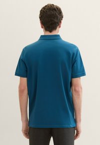 Tölgyzöld polo felső, rövid ujjú, klasszikus gallérral, sima textúrával, letisztult designnal és varrási részletekkel az alján és az ujjakon.