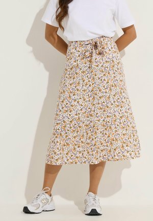 Kobieta ubrana w białą koszulkę, kwiecistą spódnicę midi z pomarańczowymi i fioletowymi kwiatami oraz białe buty sportowe New Balance, stojąca na jednolitym tle.