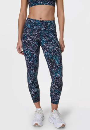 Vrouw draagt hooggetailleerde, meerkleurige legging met patroon en witte sportschoenen tegen een effen achtergrond.
