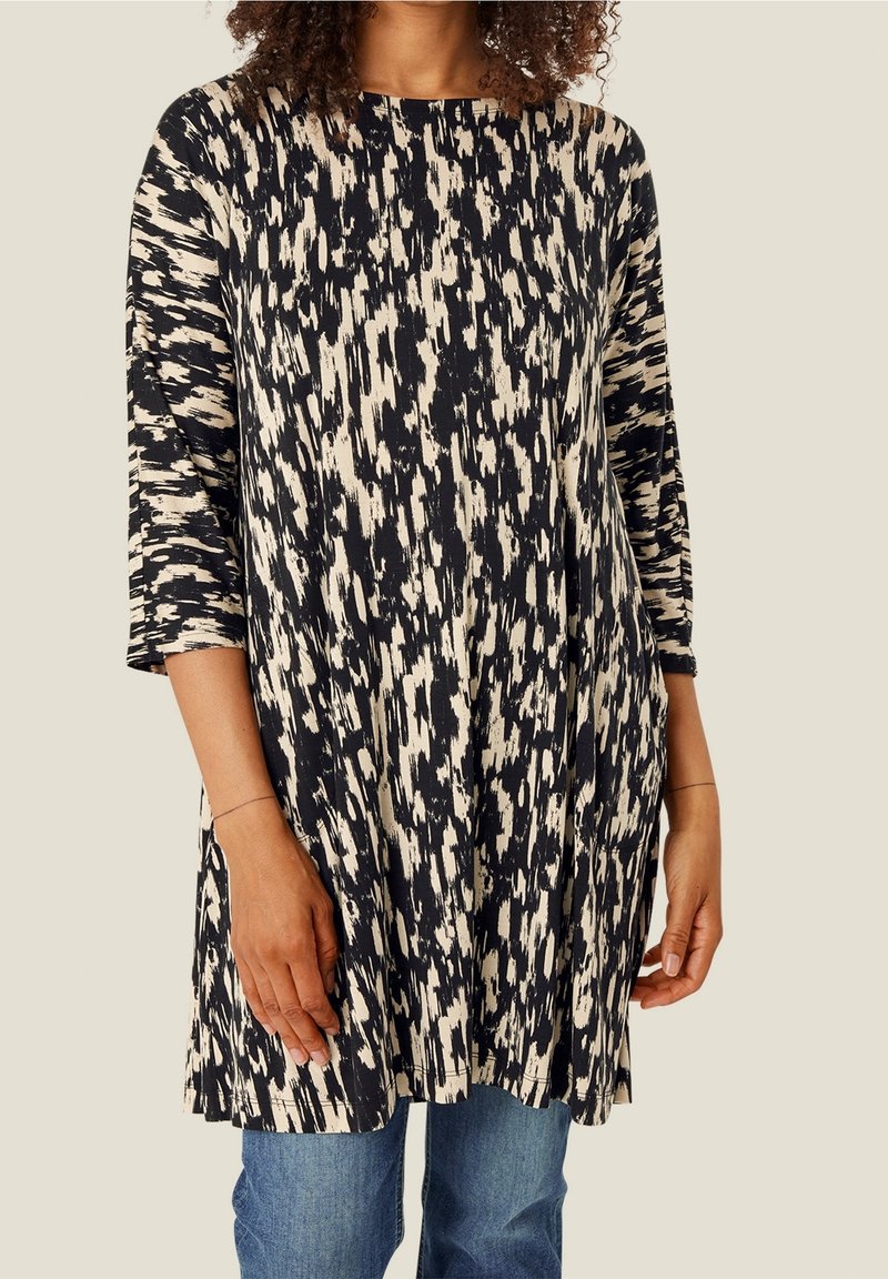 Femme portant une tunique à manches longues noire et beige à motif abstrait sur un jean bleu, debout devant un fond neutre.