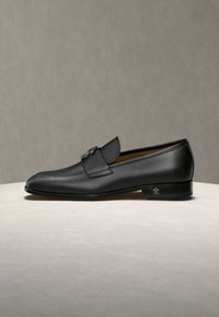 Čierna kožená pánska mokasína so strieborným detailom pracky, nízkym podpätkom a hladkým povrchom na neutrálnom podklade a pozadí.