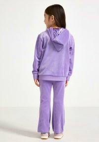 Purple velour tracksuit featuring a hooded sweatshirt and flared pants. Smooth texture with ribbed cuffs and waistband. Sneakers complete the outfit.