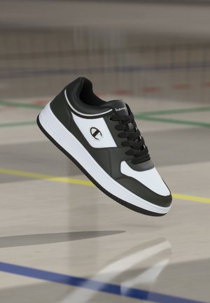 Sneaker alb-negru cu șireturi, cu logo Champion pe lateral, fotografiat în aer deasupra podelei unei săli de sport cu linii colorate pe teren.