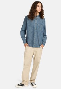 Donkerblauwe denim overhemd met knopen en een borstzak, gecombineerd met lichtbeige broek en zwarte sneakers, met een ontspannen pasvorm.
