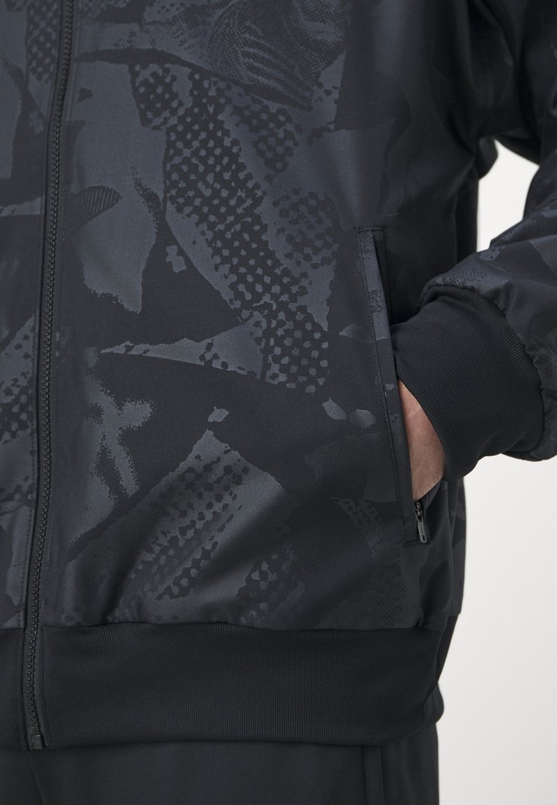 Giacca nera con zip caratterizzata da un motivo scuro texturizzato. Presenta polsini e orlo a costine, con una tasca laterale in vita.