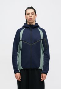 Corta-vento azul-marinho e verde claro com capuz, apresentando painéis angulares, textura suave e um bolso com fecho éclair no braço esquerdo.