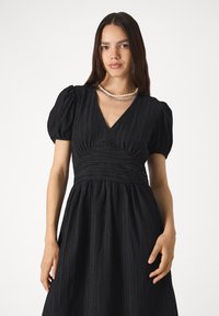 Μαύρο φόρεμα με υφή υφάσματος, φουσκωτά μανίκια και V-λαιμόκοψη. Η ρυτίδωση στη μέση και η ρέουσα φούστα ολοκληρώνουν το σχέδιο.