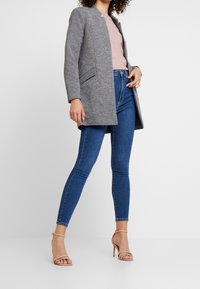 Harmaa räätälöity takki vaaleanpunaisen neuleen päällä, yhdistetty sinisiin skinny-farkkuihin ja nudenvärisiin remmisandaaleihin. Sileä kangas ja vartalonmyötäinen siluetti.