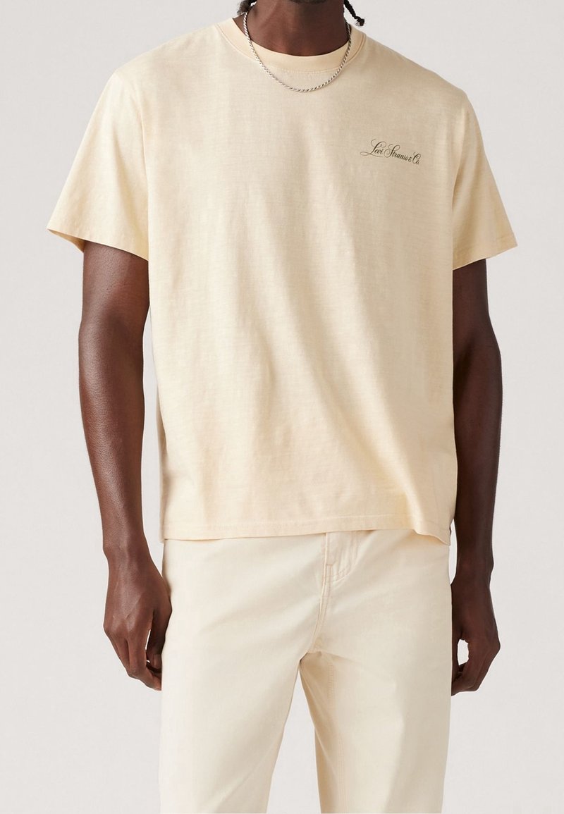 Homme portant un t-shirt beige clair à manches courtes avec un petit texte "Levi Strauss & Co." sur la poitrine et un pantalon beige clair assorti.