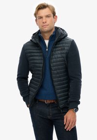 Zwart gewatteerd vest met een ritssluiting aan de voorkant, capuchon en contrasterende mouwen. Draagt over een blauwe trui en gecombineerd met donkere denim jeans.