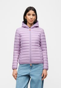 Γυναίκα που φοράει ένα ανοιχτό μοβ καπιτονέ μπουφάν με κουκούλα πάνω από ένα σκούρο πουκάμισο και χαλαρά ανοιχτό μπλε τζιν, στέκεται μπροστά σε απλό φόντο.