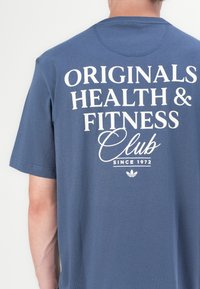 Blå t-shirt med korte ærmer lavet af bomuld, med hvid tekst: "ORIGINALS HEALTH & FITNESS Club SINCE 1972" og et adidas-logo.