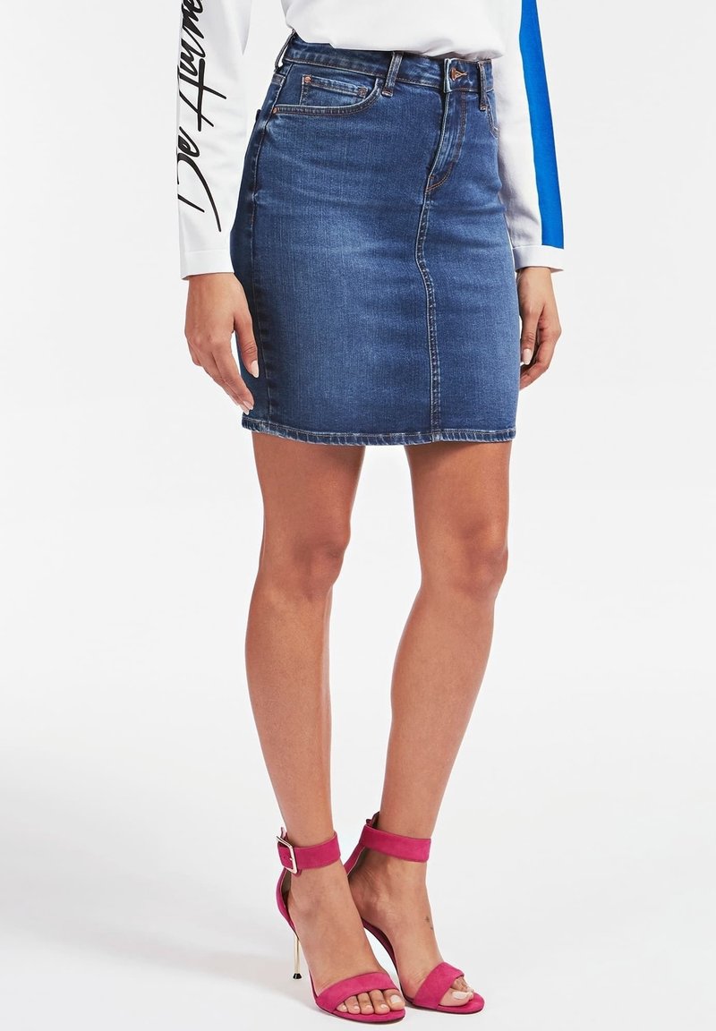 Femme portant une jupe en jean bleue, une chemise blanche à manches longues avec un texte noir sur la manche, et des sandales à talons hauts roses avec une bride à la cheville.