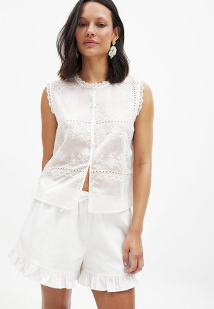 Blusa bianca senza maniche ricamata con pizzo e chiusura a bottoni, abbinata a pantaloni corti bianchi con volant in un tessuto leggero.