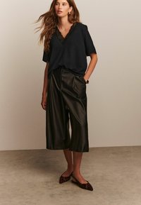 Czarna bluzka w stylu wrap z koronkowym wykończeniem, w zestawie z luźnymi czarnymi skórzanymi kulotami i wzorzystymi balerinami. Gładkie tekstury i luźny krój.