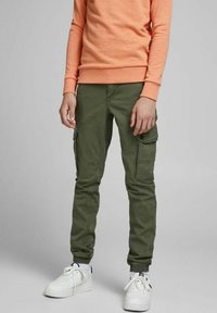 Pantaloni cargo verde oliva con molte tasche laterali, vestibilità slim, gambe affusolate e una leggera elasticità. Abbinati a sneaker bianche e una felpa arancione.