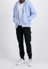 Sudadera con capucha y cremallera en azul claro con cordones, usada sobre una camiseta blanca. Pantalones cargo negros con varios bolsillos y puños elásticos, combinados con zapatillas blancas.