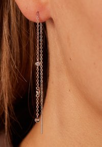 Boucles d'oreilles en chaîne argentée avec un design linéaire, comportant de petits éléments décoratifs et une barre pendante, sur un fond de teint.