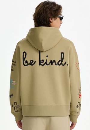 Personne portant un sweat à capuche beige avec le texte "be kind." dans le dos et divers petits motifs graphiques sur les manches, tournée vers l'arrière avec des lunettes de soleil.