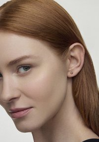 Złoty, sercowy, mały stud w uchu. Gładka powierzchnia, minimalistyczny design, podkreślający naturalny odcień skóry.