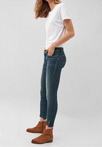 Jeans ajustados en denim azul oscuro, con cinco bolsillos, un desgastado sutil en los muslos, combinados con botas de ante marrón hasta el tobillo con acentos en zigzag.