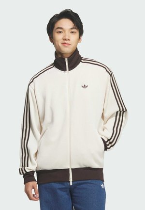 Młody mężczyzna stojący, ubrany w białą i brązową kurtkę Adidas na suwak oraz niebieskie spodnie Adidas, ręce w kieszeniach, neutralne tło.