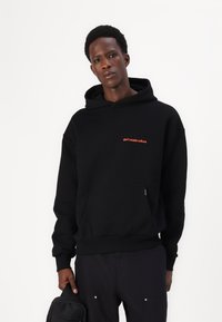 Schwarzer Kapuzenpullover mit einer Fronttasche und rot gesticktem Text "nicht Kultur verschwenden", aus strukturiertem Stoff gefertigt.