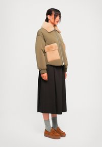Donna che indossa un giaccone a collo alto olivastro con colletto in pelliccia sintetica beige e tasche, gonna midi nera, calze grigie e mocassini con zeppa marroni.