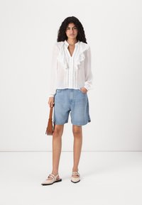 Weißes Langarmbluse mit Rüschendetails, kombiniert mit hellblauen Denim-Shorts. Das Model trägt eine braune Umhängetasche und hellfarbige Schuhe.