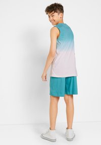 Ragazzo che sorride sopra la spalla, indossando una maglietta senza maniche in gradiente verde acqua e rosa, pantaloncini teal e scarpe alte bianche, ritratto su sfondo bianco.