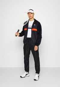 Cazadora negra con acentos naranjas, camiseta blanca con logo, pantalones de chándal negros, sosteniendo una raqueta de tenis, usando una visera blanca y zapatillas.