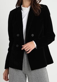 Černý sametový dvouřadý blazer s klopami a čtyřmi kulatými černými knoflíky, nošený přes bílý top s vysokým límcem a vzorované kalhoty.