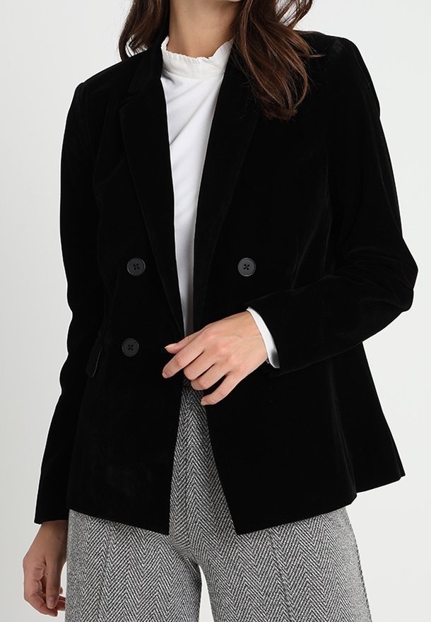 Černý sametový dvouřadý blazer s klopami a čtyřmi kulatými černými knoflíky, nošený přes bílý top s vysokým límcem a vzorované kalhoty.