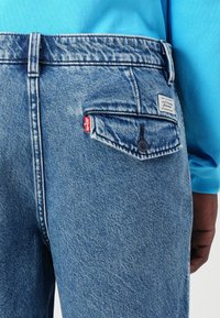 Jeans de mezclilla azul claro con un acabado desgastado, que presentan un bolsillo trasero con una etiqueta roja de Levi's y un pequeño parche de la marca, con costuras resistentes.
