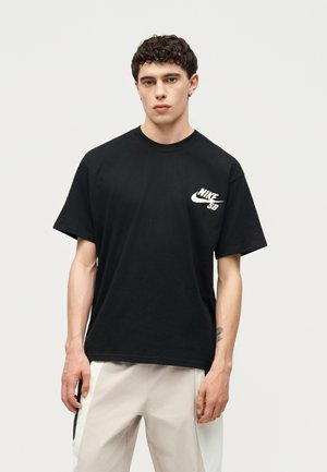 Jonge man staand, draagt een zwart Nike SB t-shirt en lichtgekleurde broek met een neutrale uitdrukking op een effen achtergrond.