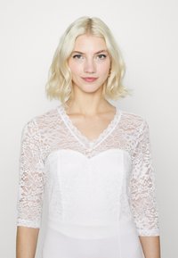 Vestido de corpete de renda branca com mangas três quartos, decote em V e padrões florais na renda, com parte inferior em tecido branco liso.