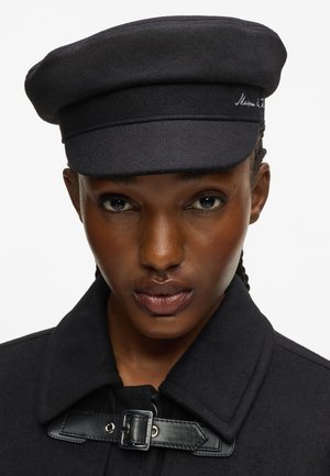 Kvinde med mørk hud iført en sort uldnyhedshat og en sort frakke med læderspændebånd, vendt fremad mod en hvid baggrund.