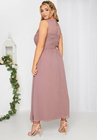 Maksymalna sukienka w kolorze mauve, z teksturowanym gorsetem ozdobionym kwiatowymi akcentami, gładką, swobodnie opadającą spódnicą oraz detalu z kluczowym otworem na plecach. Noszona z cielistymi obcasami.