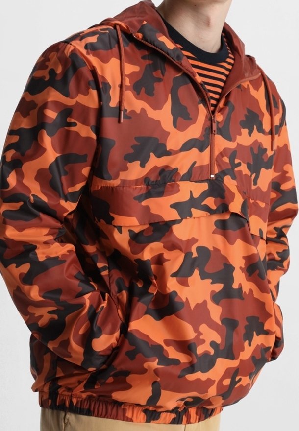 Personne portant une veste à capuche orange et noire avec un motif camouflage, une poche avant et un col zippé.