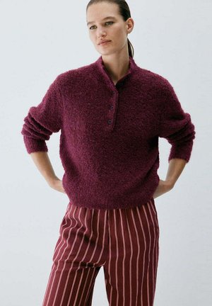 Mujer con suéter abotonado de textura granate y pantalones granates con finas rayas verticales blancas, de pie con las manos en las caderas.