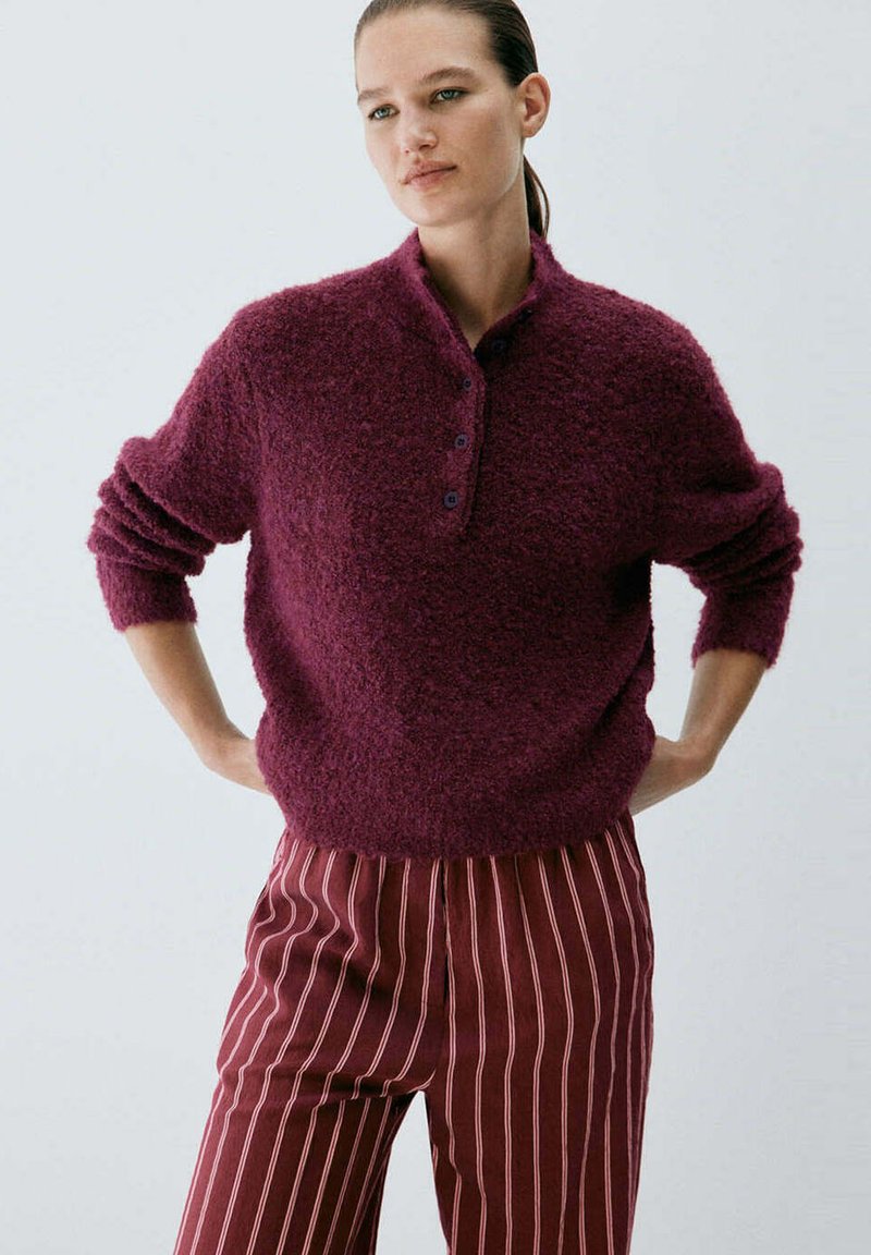 Femme portant un pull texturé bordeaux à boutons et un pantalon bordeaux avec de fines rayures blanches verticales, debout, les mains sur les hanches.