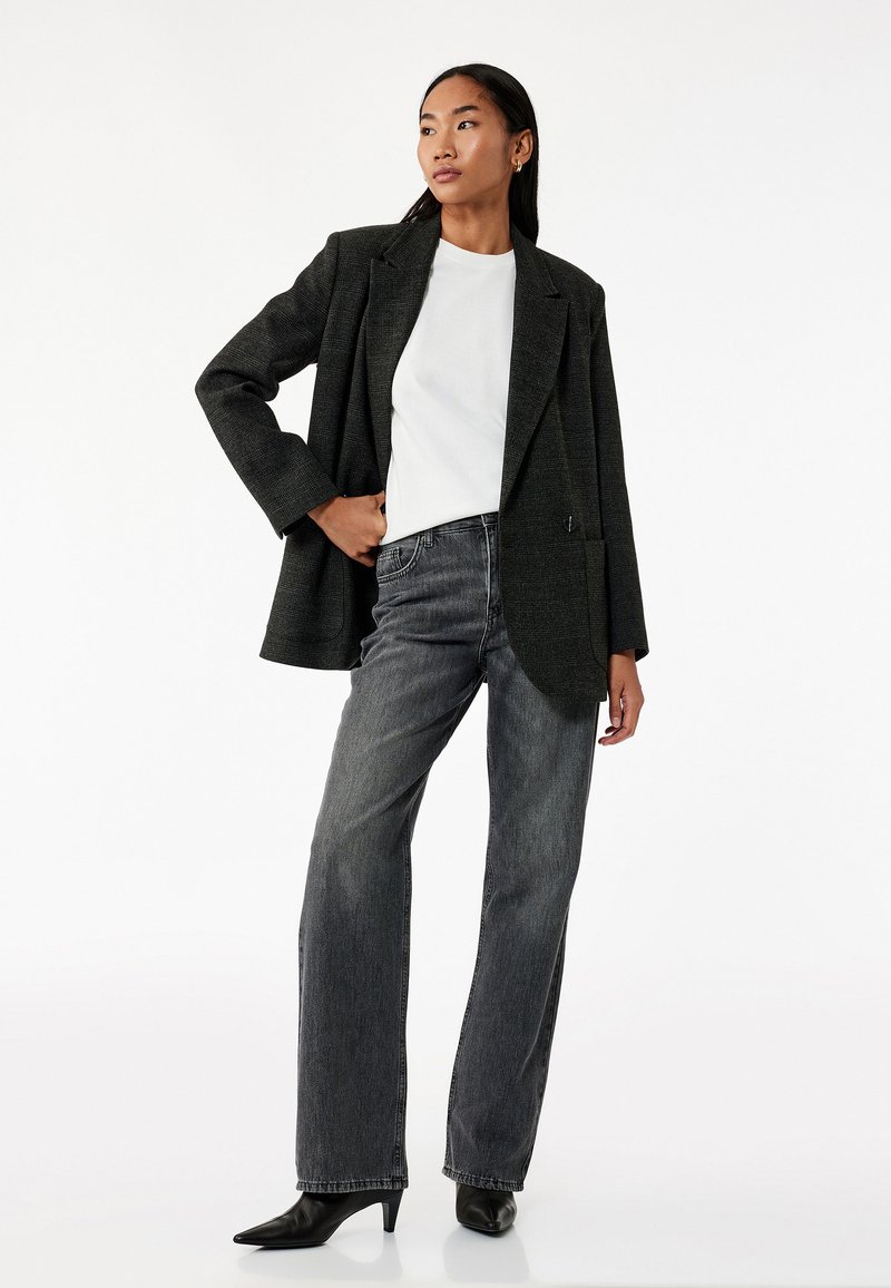 Blazer gris oscuro de tela texturizada, camiseta blanca de cuello redondo y jeans negros de pierna recta. El modelo lleva tacones negros puntiagudos.