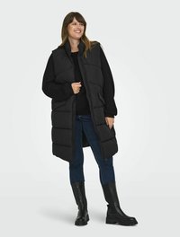 Svart, ärmlös pufferväst med hög krage, bärs över en svart tröja. Matchas med mörka jeans och knähöga svarta stövlar.