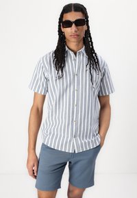Chemise à manches courtes à boutons avec des rayures verticales bleues et blanches, accompagnée d'un short bleu. La chemise a un col et des boutons beiges.