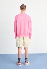 Rosa Sweatshirt mit gerippten Bündchen und Saum, kombiniert mit beigen Shorts. Socken mit Camouflage-Muster und rosa Sneakers mit weißen Akzenten.