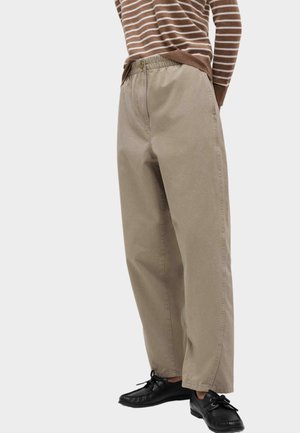 Person iført beige vidde bukser med elastisk talje, brun- og hvide stribede sweater og sorte snørestøvler stående.